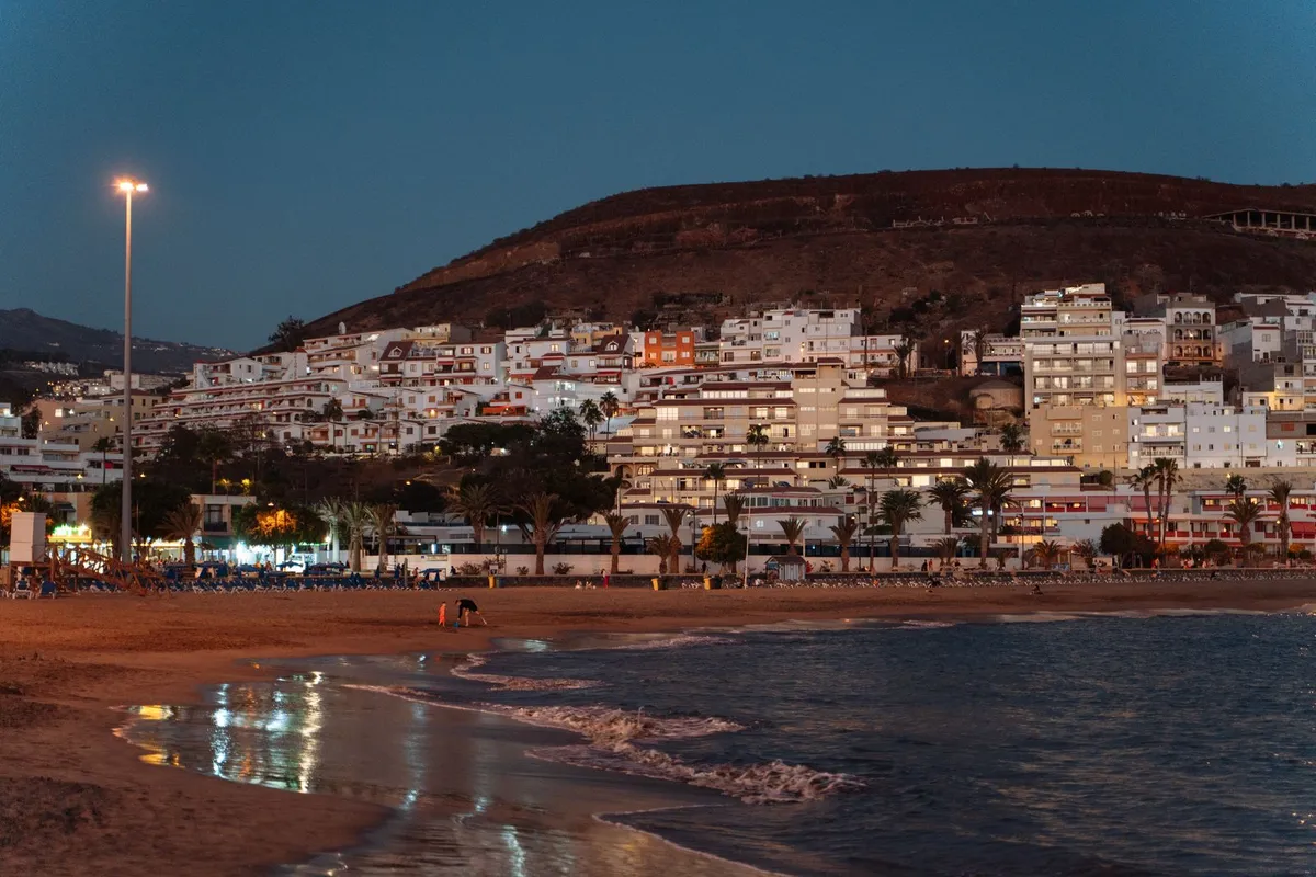 A south Tenerife town beach at dusk, with lit white apartment terraces rising up the hillside behind the sand