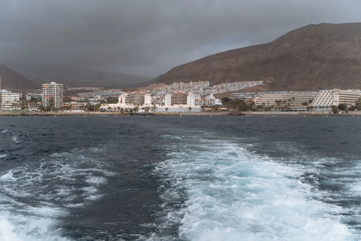 A south-coast resort town seen from a departing boat, with a wake trailing across the water and a bare volcanic hill rising behind the buildings