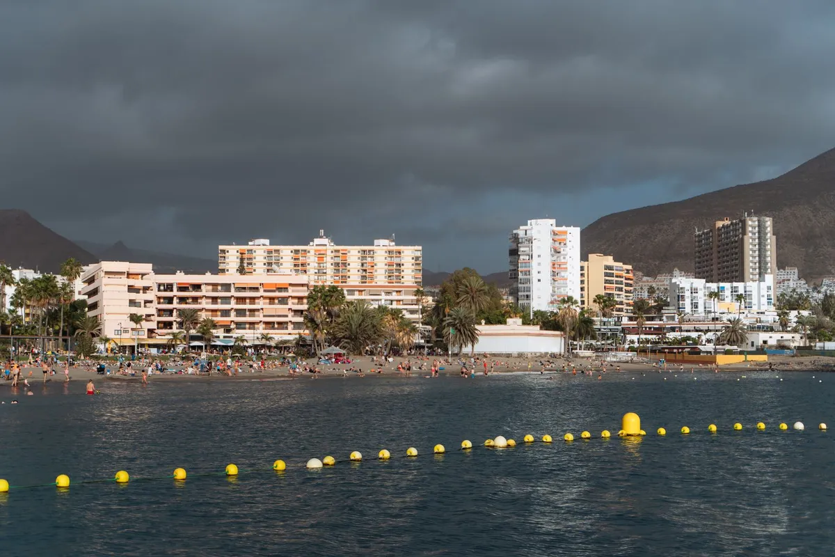A busy golden-sand town beach with a yellow buoy line marking the swimming zone, backed by hotel blocks and mountains