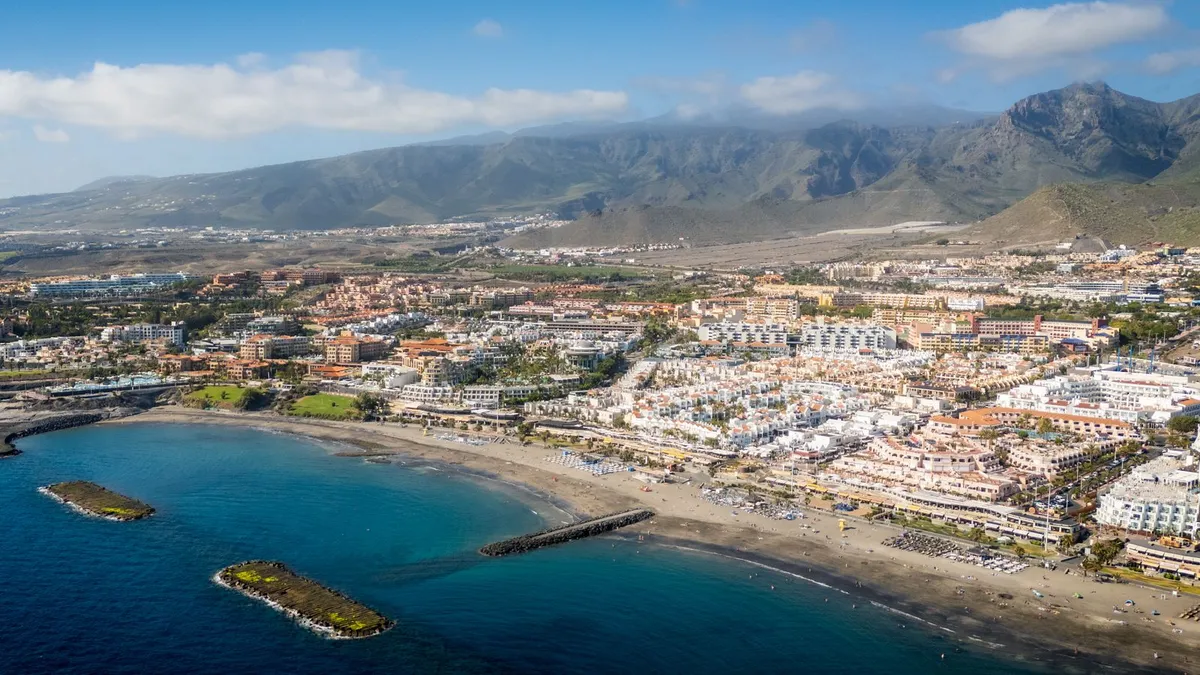 Aerial view of the south Tenerife resort coast, with dense hotels, breakwater-sheltered beaches and mountains behind