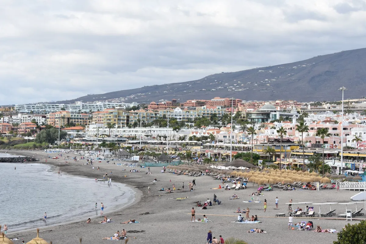 Sunbathers and loungers on a golden-sand, breakwater-sheltered beach backed by resort hotels and mountains on the south Tenerife coast